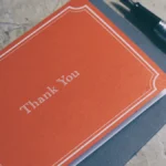 Orange thank you note to nurse staff resting on a grey envelope beside a silver fountain pen